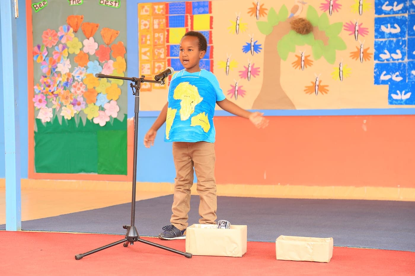 A boy speaking into a microphone with his arms open, wearing a hand-painted world map T-shirt